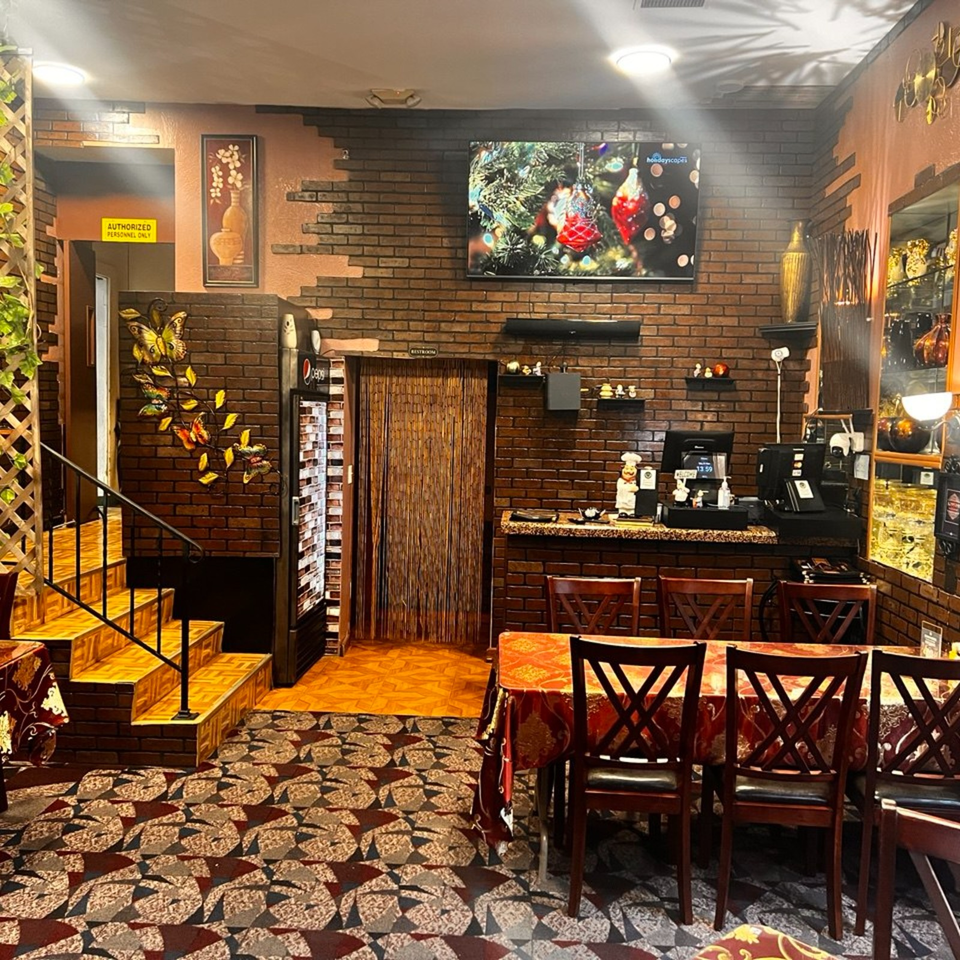 Warmly lit restaurant interior with brick walls, patterned flooring, dining tables, reception counter, staircase, and decorative displays.