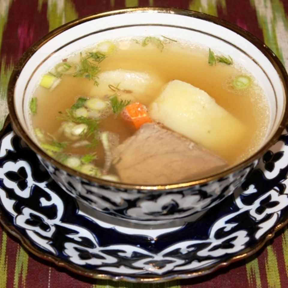 Bowl of clear broth soup with potatoes, meat, carrots, and fresh herbs on an ornate patterned plate.