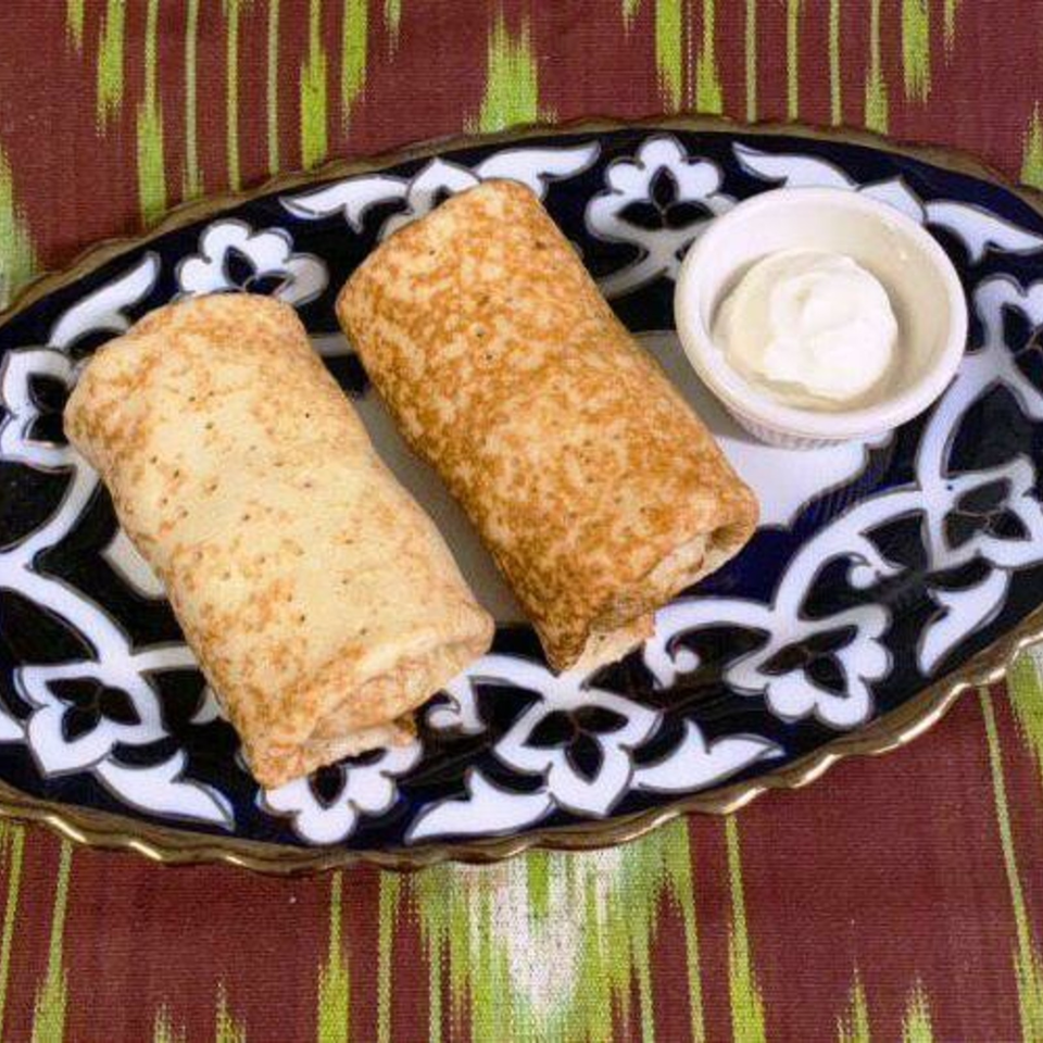 Two golden rolled crepes served with a bowl of creamy white dipping sauce on an ornate black-and-white plate.