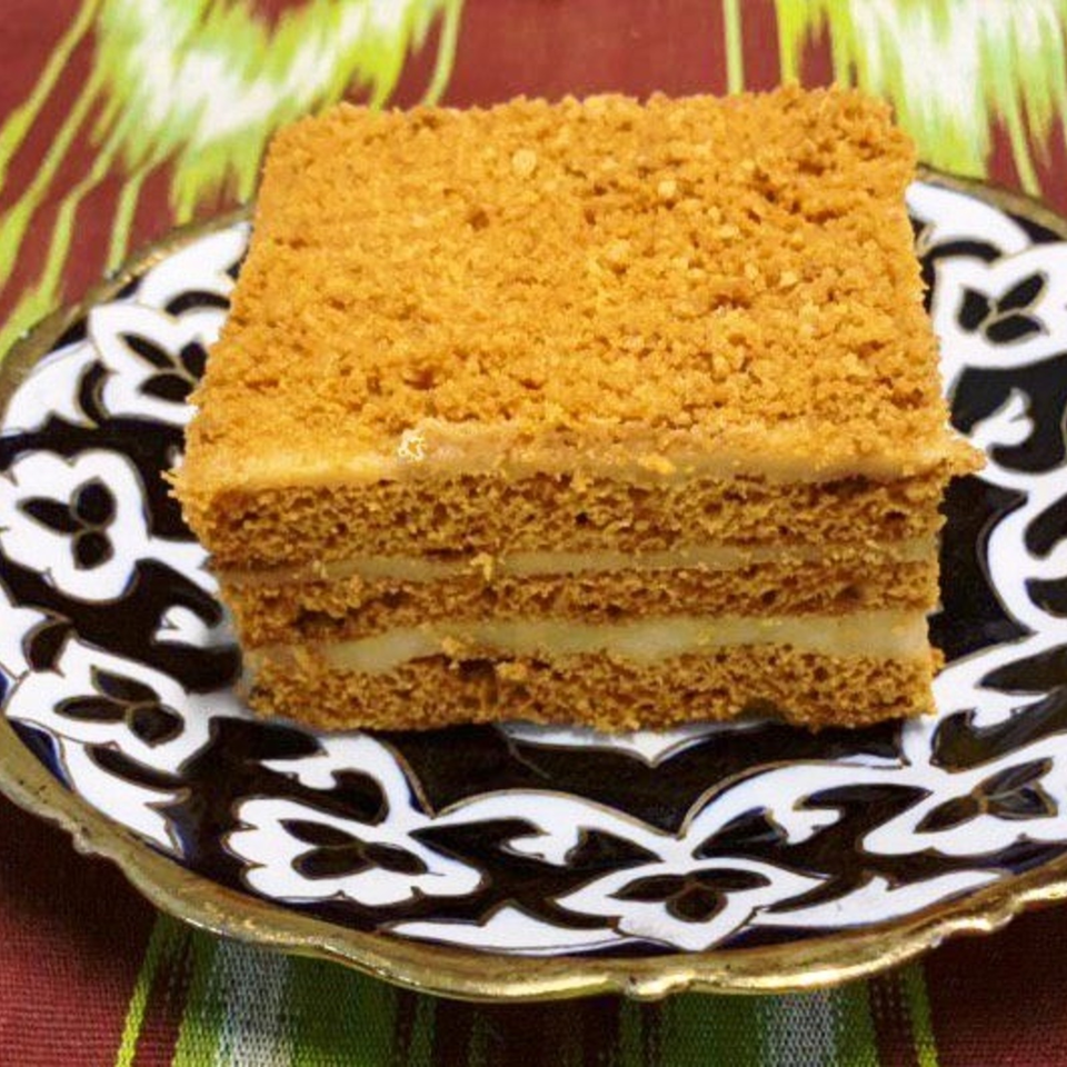 Square slice of layered golden cake with crumb topping and pale filling, served on an ornate black-and-white patterned plate.