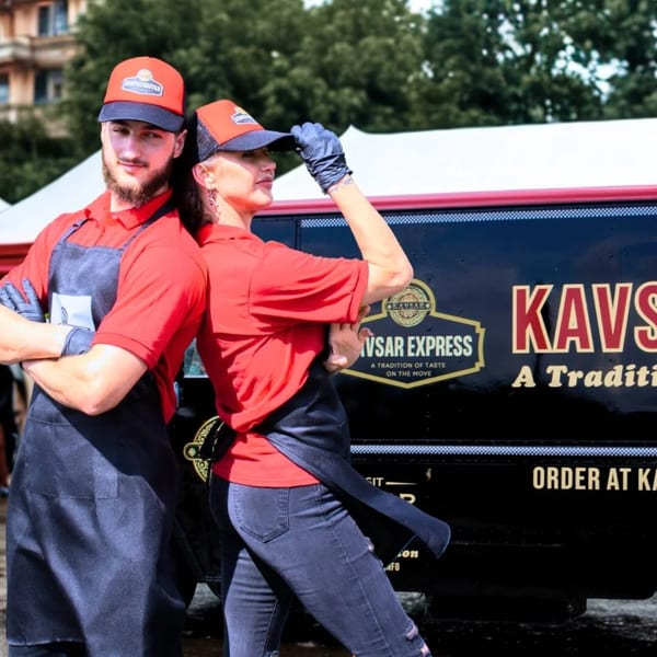 Two Kavsar Restaurant staff in red shirts and aprons pose next to a black food truck.
