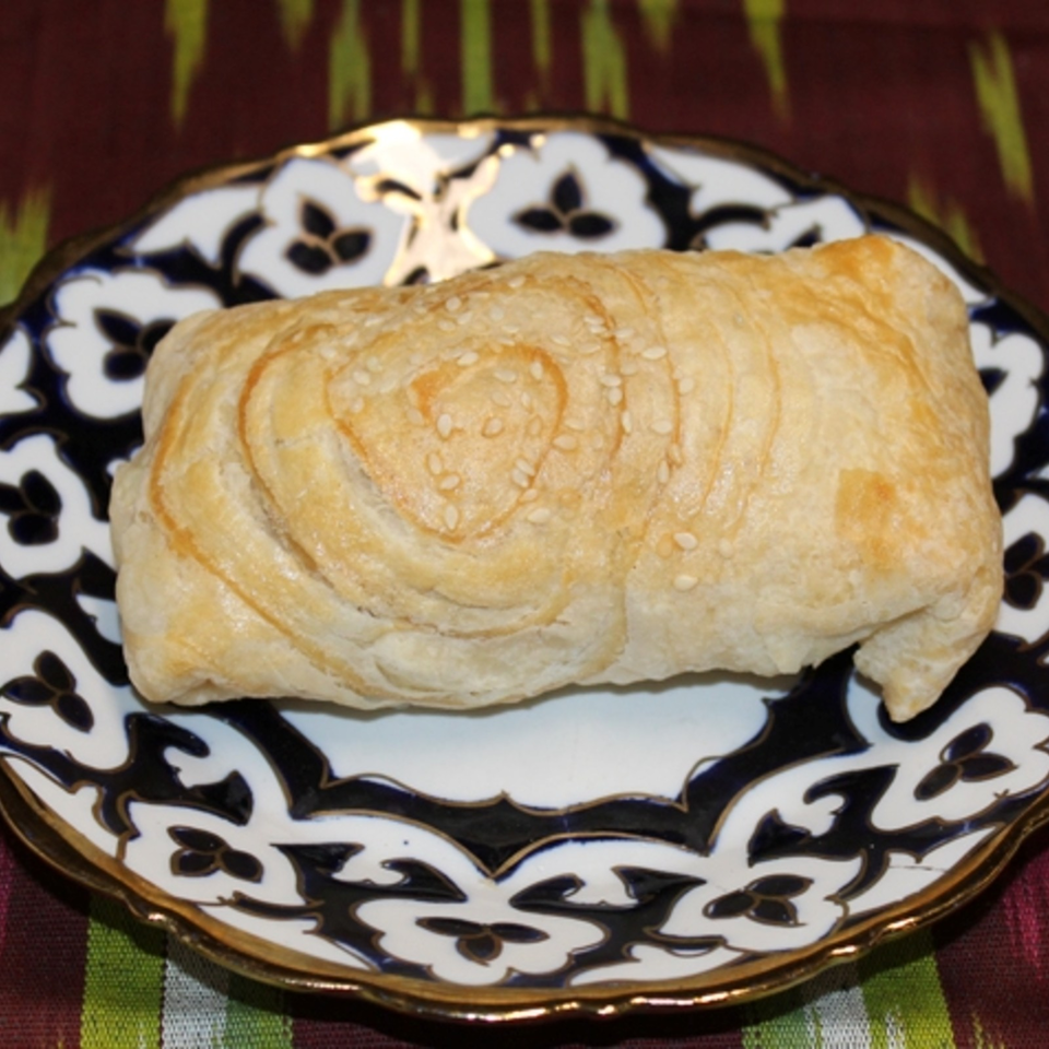 Golden sesame-topped flaky pastry served on an ornate black-and-white decorative plate.