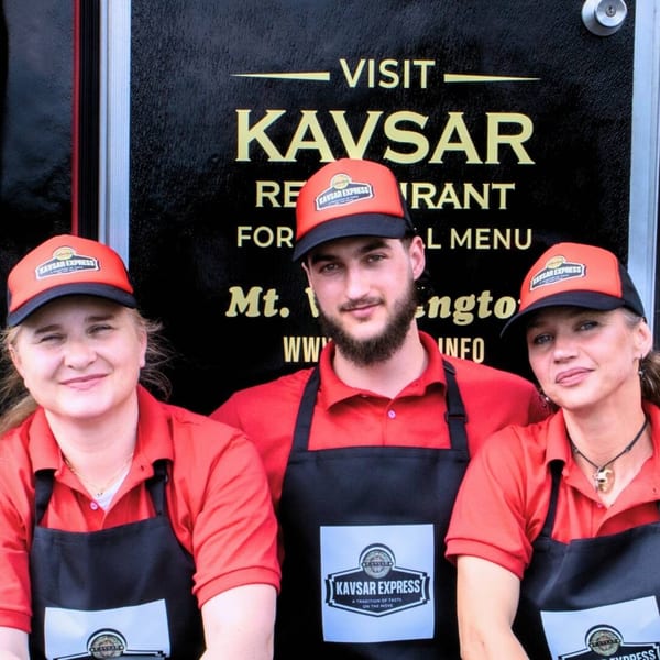 Three Kavsar Express staff members in red shirts and black aprons smiling in front of a food truck.