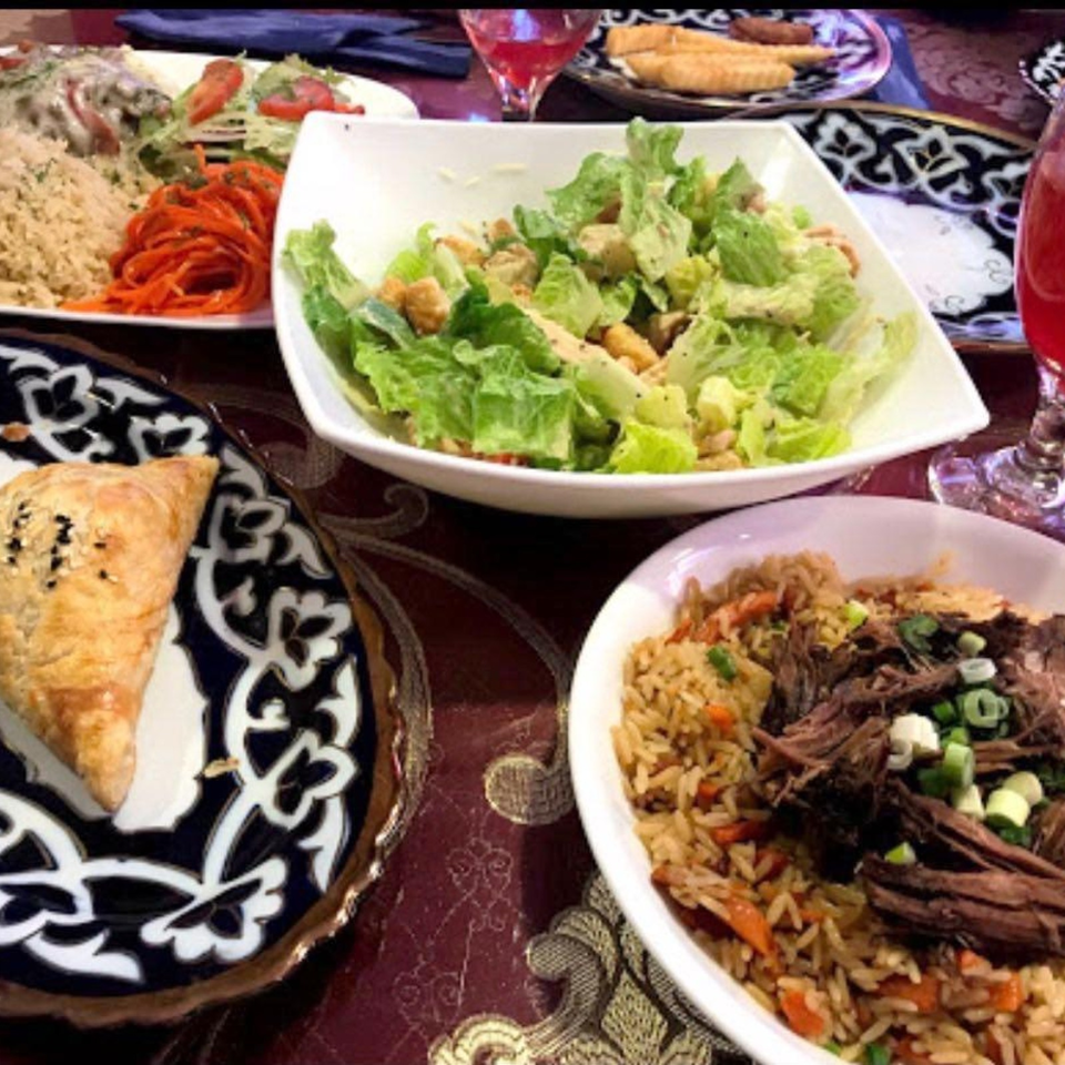 Middle Eastern-style spread featuring green salad, spiced rice with shredded meat, pastries, vegetables, and red drinks on patterned tablecloth.