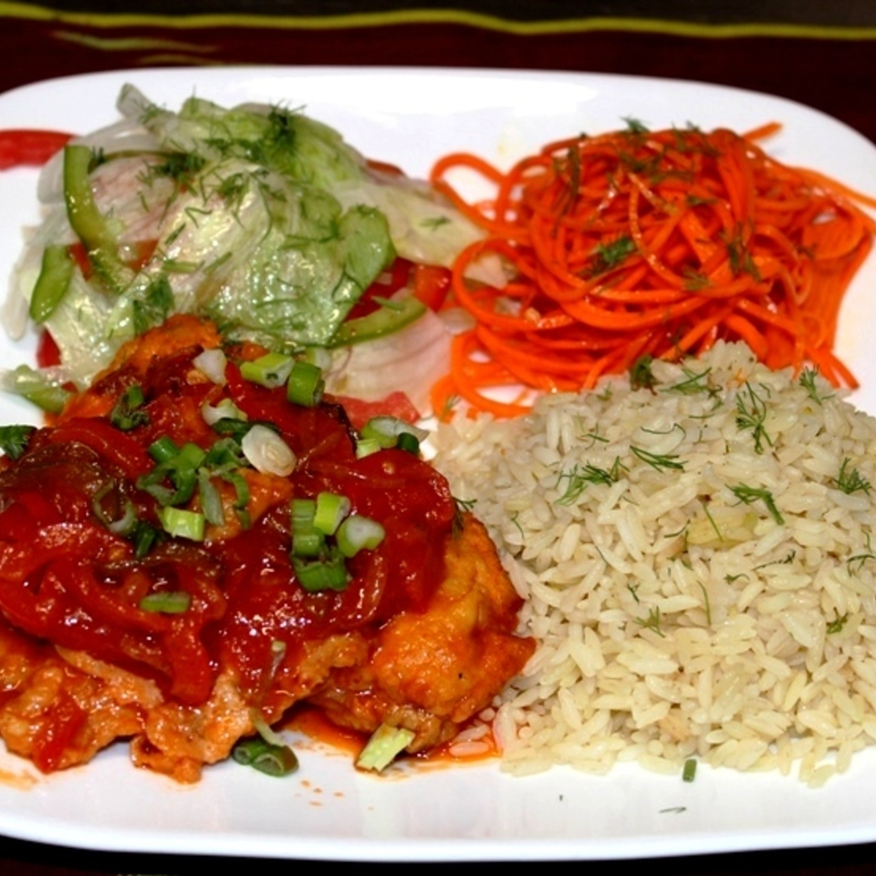 Plate of rice, tomato-topped fried chicken, shredded carrot salad, and cucumber salad garnished with fresh herbs.