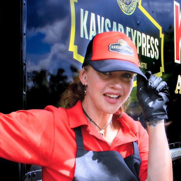 Smiling Kavsar Restaurant employee in a red shirt, black apron, and branded cap, against a food truck.