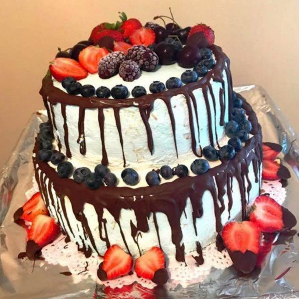 Two-tier white cake with chocolate ganache drips, blueberries, strawberries, blackberries, and cherries on top and around the base.