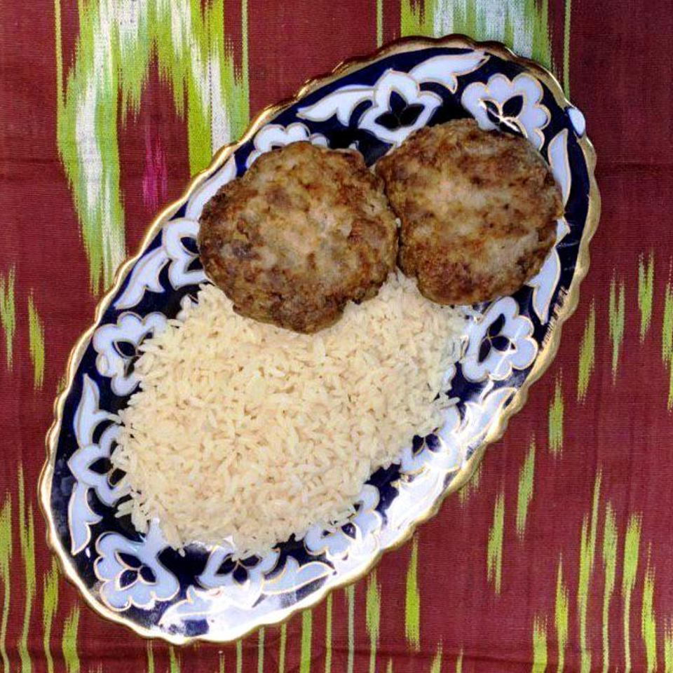 Two browned meat patties served with white rice on an ornate blue-and-white platter atop a striped tablecloth.