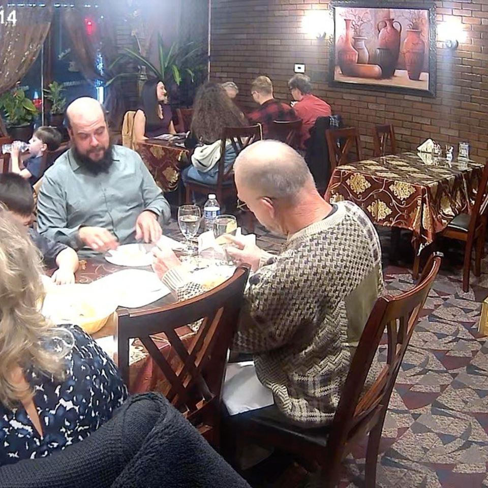 Busy restaurant dining room with several groups eating at patterned tables, surrounded by brick walls, plants, and ceramic artwork.