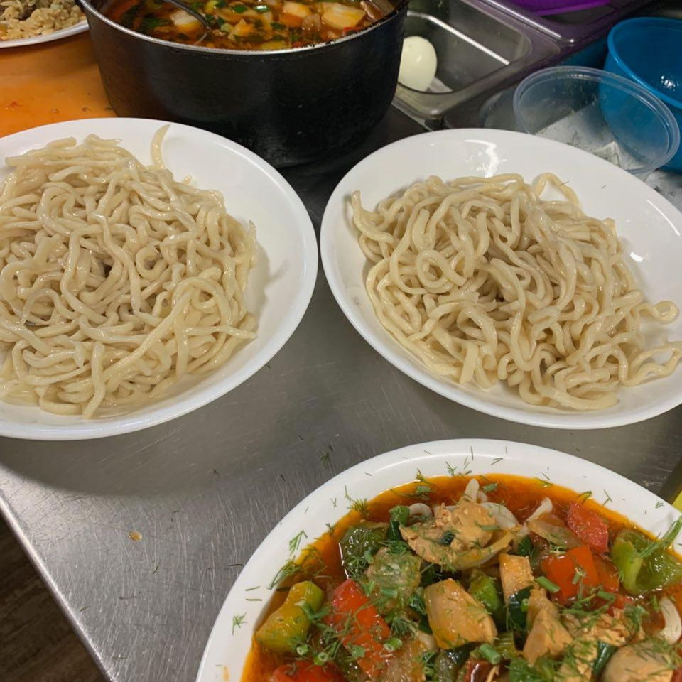 Two plates of fresh noodles sit beside a bowl of colorful vegetable and meat stew on a commercial kitchen counter.