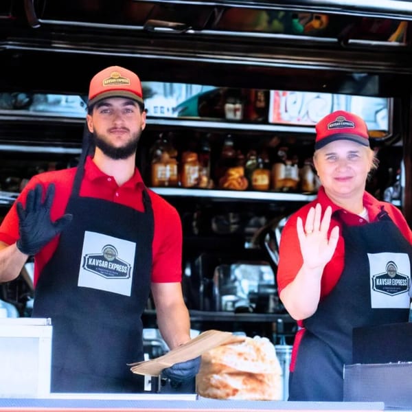 Two Kavsar Express employees, a man and a woman, in red shirts and black aprons, waving.