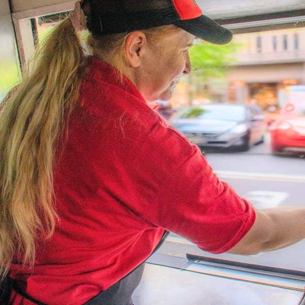 A woman in a red shirt and black cap looks out from a food truck window at a street.