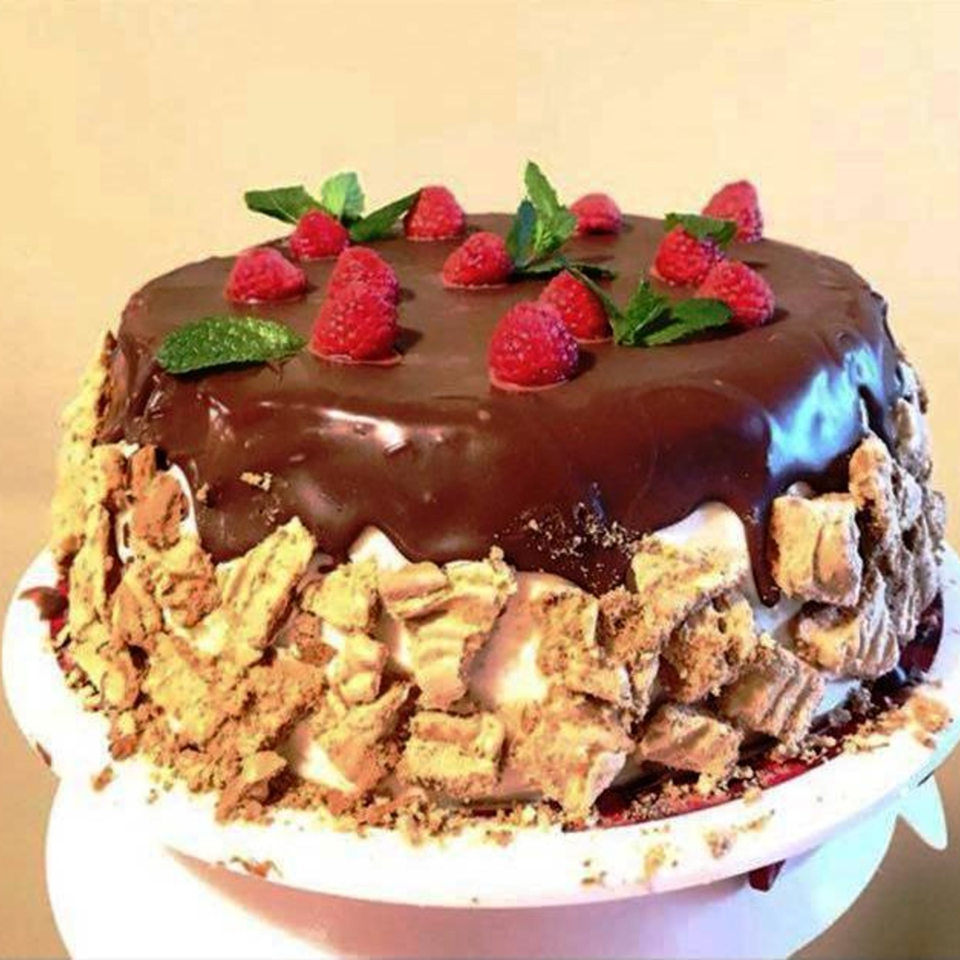 Chocolate-glazed cake covered with raspberries, mint leaves, and crumbled pastry on a white plate.